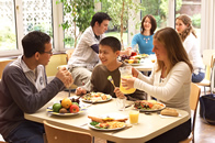 Students in small groups seated at tables having lunch in sun-lit dining hall, with windows showing green of garden beyond