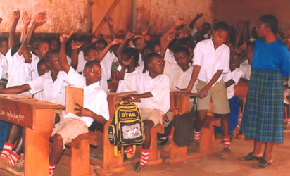 classroom of teacher and children, the children all with hands raised to answer a question