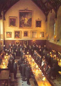 Atmospheric image of the Dining Hall in Exeter College, with diners in black robes standing at long tables before eating, and a golden light from the many small lamps on the tables giving the hall a nice ambience